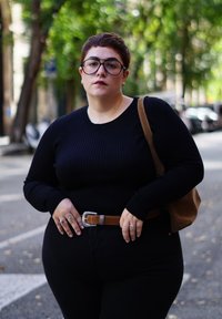 Cilvēks ar īsiem matiem un brillēm, valkājot melnu džemperi un jostu, stāv uz koku ieskautas ielas ar brūnu pleca somu.