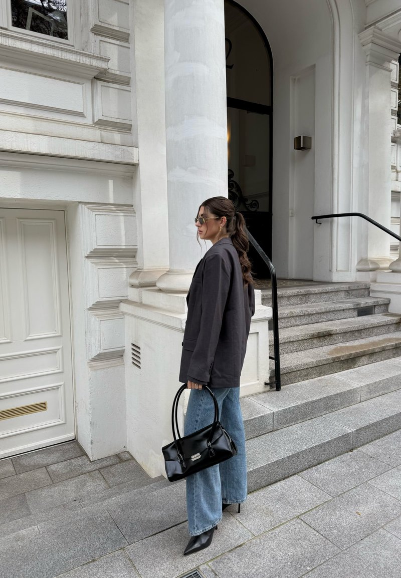 Bolso negro con asas dobles, combinado con un blazer oscuro y jeans de pierna ancha azules. El escenario presenta un edificio blanco y pasos de piedra gris.
