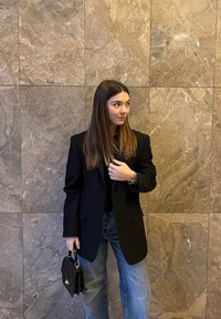 Czarna dopasowana marynarka na czarnym topie, zestawiona z jasnoniebieskimi szerokimi jeansami, trzyma małą czarną torebkę, na tle teksturowanej kamiennej ściany.