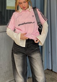 Roza majica Adidas s črnim besedilom in zvezdami, ki je nameščena čez belo majico z dolgimi rokavi. Udobno oblečena z raztrganimi širokimi črnimi jeans hlačami.
