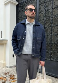 Mężczyzna ubrany w ciemnoniebieską kurtkę dżinsową, szary sweter, szare spodnie i okulary przeciwsłoneczne, trzymający beżową torbę na ramię przy czarnej dekoracyjnej bramie na świeżym powietrzu.