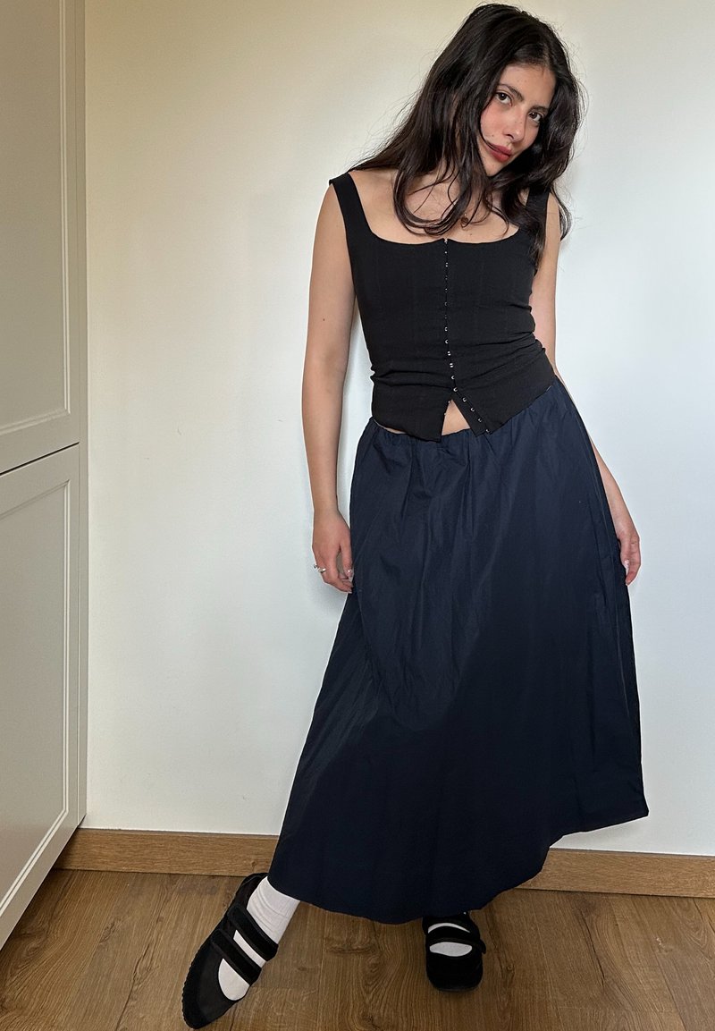 Femme aux longs cheveux foncés portant un haut noir sans manches, une longue jupe marine, des chaussettes blanches et des chaussures Mary Jane noires, debout à l'intérieur sur un sol en bois.