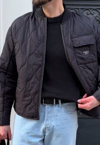 Černá prošívaná bomber bunda se zipem vpředu, stojáčkovým límcem a přední kapsou, nošená přes černý svetr a světle modré džíny.