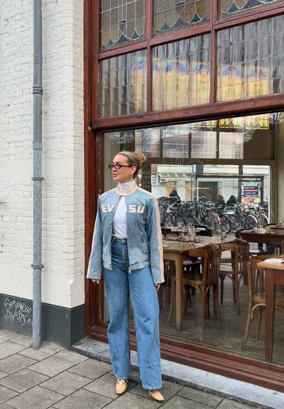 Chaqueta de mezclilla con acentos beiges y letras "EVISU", suéter de cuello alto blanco, vaqueros de pierna ancha azules y zapatos en tonos neutros frente a un restaurante.