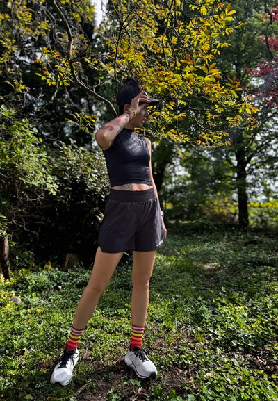 Top corto atlético negro y pantalones cortos, calcetines a rayas multicolores, zapatillas de running blancas, de pie al aire libre entre el follaje verde y las hojas amarillas.