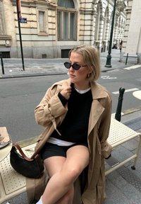 Trench-coat beige porté sur un pull noir, assorti à un short foncé. Accessoires : des lunettes de soleil et un sac à main marron sur un banc perforé.