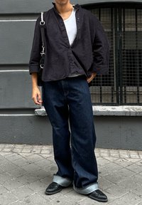 Persona che indossa jeans scuri larghi con risvolti, scarpe nere, T-shirt bianca, camicia nera a bottoni aperta e porta una borsa a tracolla nera mentre si trova sul marciapiede.
