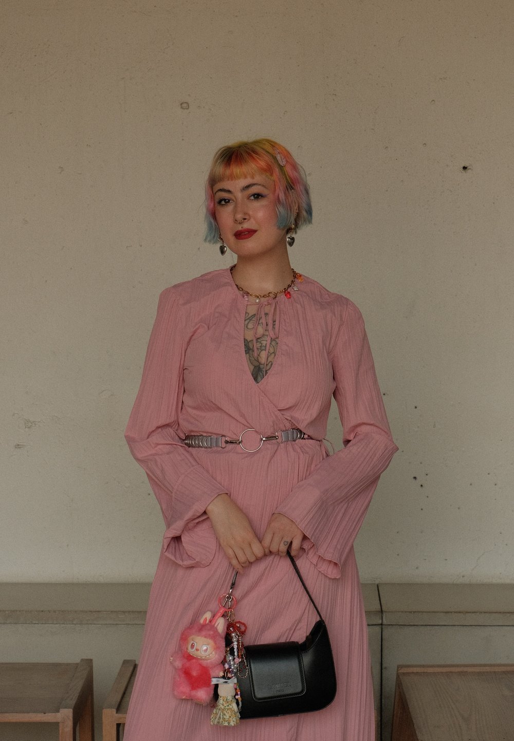 Vestido envolvente rosa con mangas largas, tejido texturizado y un cinturón plateado como acento. Bolsa negra con charms, que incluyen un conejito de peluche rosa y un llavero.