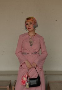 Vestido envolvente rosa con mangas largas, tejido texturizado y un cinturón plateado como acento. Bolsa negra con charms, que incluyen un conejito de peluche rosa y un llavero.