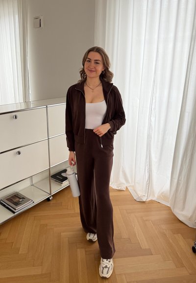 Chándal de terciopelo marrón con chaqueta con cremallera y pantalones de pierna ancha, que lleva sobre una camiseta blanca. Sostiene una botella de agua gris, de pie sobre un suelo de madera.