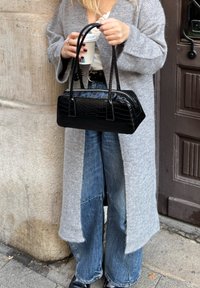 Čierna kabelka s krokodílím vzorom a dvojitými rúčkami, držená osobou v sivom kabáte a modrých širokých džínsach s kávovým pohárom v druhej ruke.