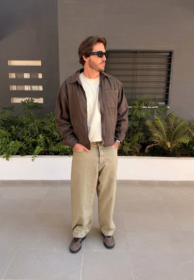 Hombre con gafas de sol, chaqueta marrón, camisa blanca y pantalones beige, de pie con las manos en los bolsillos frente a plantas y una pared gris.