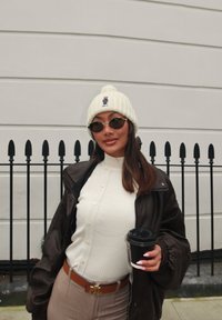 Jaqueta de pele castanha na moda, suéter creme de relevo, calças bege, gorro branco com detalhe, óculos de sol redondos, segurando uma chávena de café preto.