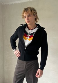 Veste noire zippée avec une texture côtelée portée sur une chemise  à motifs multicolores comportant des chevrons rouges, jaunes et noirs, complétée par un pantalon à carreaux.