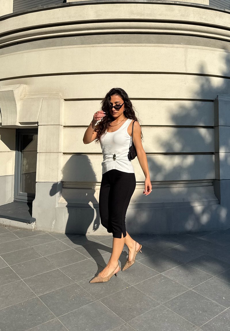 Γυναίκα με γυαλιά ηλίου, που φοράει λευκό αμάνικο τοπ, μαύρο κοντό παντελόνι και μπεζ ψηλοτάκουνα περπατά σε πλακόστρωτη περιοχή δίπλα σε ένα καμπυλωτό πέτρινο κτίριο.