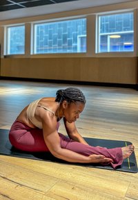 Une personne s'étire sur un tapis de yoga noir, portant un crop top beige et un pantalon ajusté bordeaux, dans un espace intérieur bien éclairé avec un sol en bois.