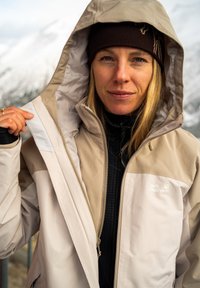 Beige ja musta ulkotakki, jossa korkea kaulus ja huppu, vetoketju ja logotunnus. Sileä, sääsuojaava kangas.