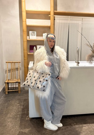 Joven mujer con sudadera gris con capucha y pantalones deportivos, con abrigo de piel blanco, de pie en cocina moderna sosteniendo una bolsa plateada con estampado de estrellas.