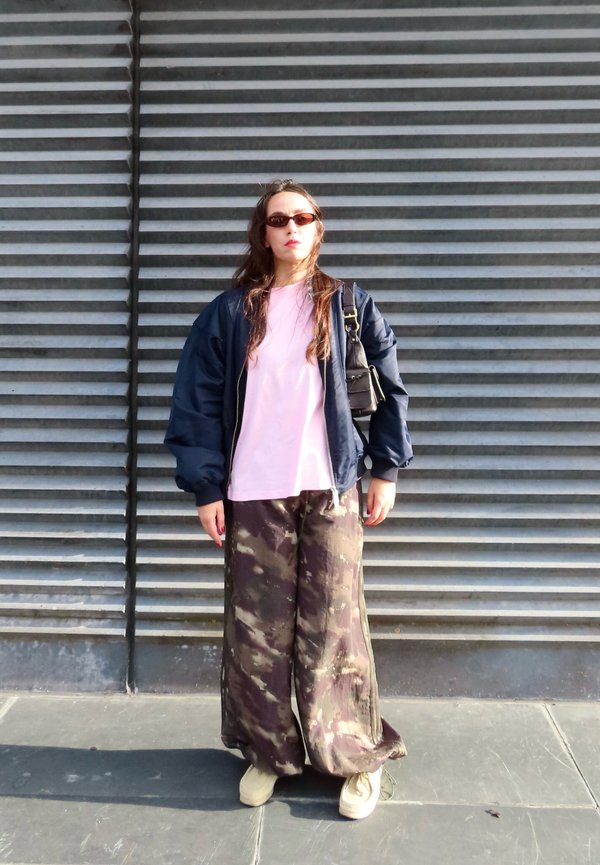 Une femme portant des lunettes de soleil, une veste bleu marine, une chemise rose, un pantalon camouflage et des chaussures blanches se tient devant un mur métallique à persiennes.