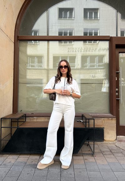 Camiseta blanca corta y pantalones de campana blancos, combinados con zapatos de plataforma beige. Accesorio incluye un pequeño bolso marrón. Entorno urbano.