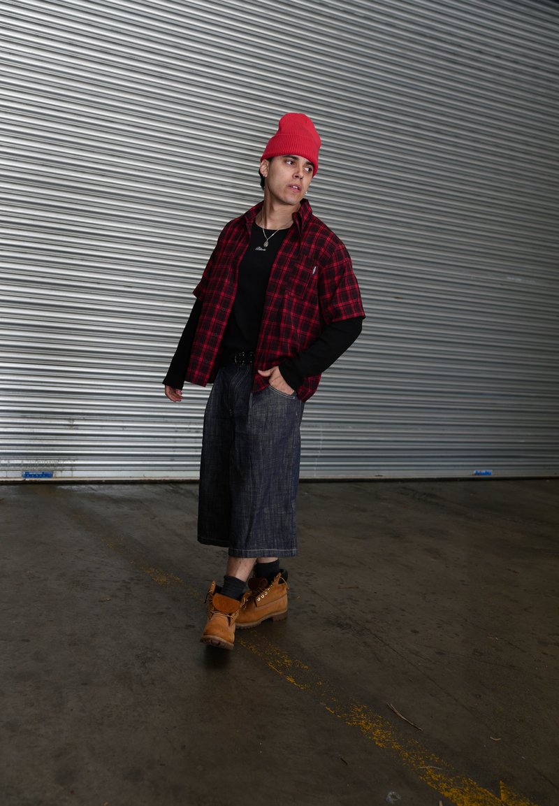 Jong persoon met een rode beanie, een rood geruit hemd over zwarte lange mouwen, brede spijkershorts en beige laarzen, wandelt binnen langs een metalen rolluik.