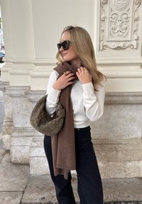 Strikket hvid sweater, brun tørklæde og mørke jeans. Tilbehøret omfatter en vævet olivengrøn taske og guldringe. Glatte teksturer og enkelt design.