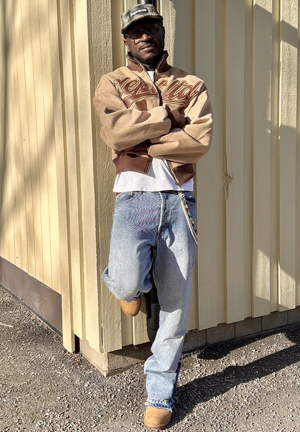 Homme portant une veste marron, une chemise blanche, un jean bleu, des bottes beige et une casquette noire Supreme, appuyé contre un mur en bois jaune, les bras croisés.