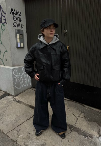 Persona con chaqueta de cuero negra, sudadera con capucha gris, gorra negra y pantalones anchos oscuros, de pie con las manos en los bolsillos sobre una acera urbana marcada con grafitis.