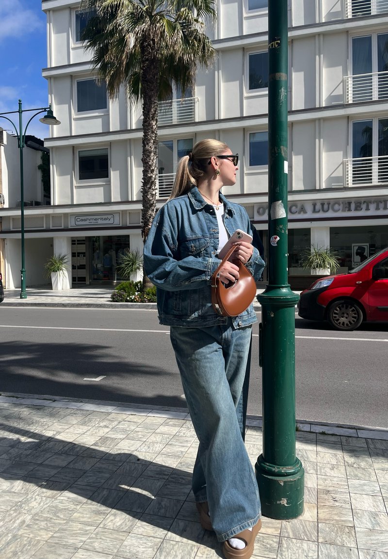 Τζάκετ από τζιν και παντελόνι με φαρδιά πόδια σε ανοιχτό μπλε, συνδυασμένα με καφέ κυκλική τσάντα. Σε φόντο μια αστική οδό με φοινικόδεντρα και σύγχρονα κτίρια.