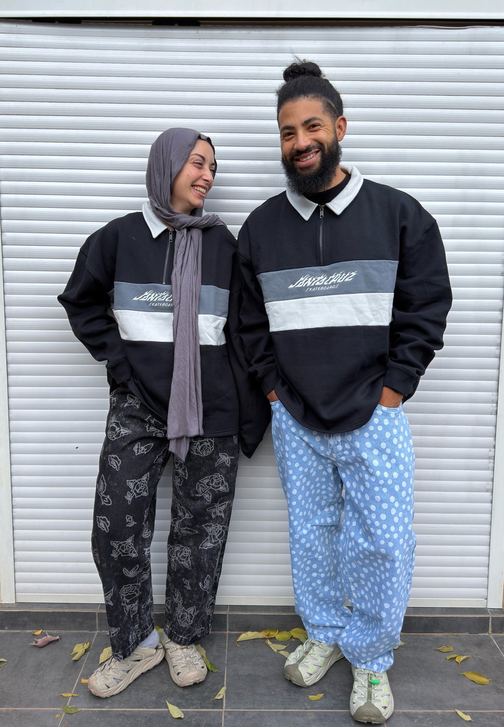 Deux personnes souriantes portant des sweat-shirts zippés assortis noirs, gris et blancs, des pantalons à motifs et des baskets debout sur un sol carrelé.