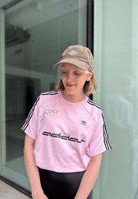 Pink atletisk skjorte med sorte striber og "adidas" logo, der har en revnet tekstur og stjerne grafik. Brun kasket med tekst og flad skygge.