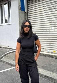 Donna con capelli scuri alla spalla, indossando un dolcevita nero e pantaloni, occhiali da sole e una borsa nera, si trova sul marciapiede urbano.