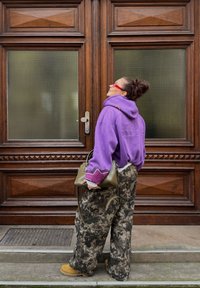 Sweat à capuche oversize violet avec des accents roses contrastants, pantalon large à motif camouflage, et un sac à bandoulière vert, associés à des bottes couleur tan.
