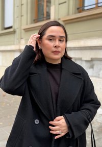 Sort dobbeltradet uldfrakke med store knapper, båret over en mørk sweater. Personen har en struktureret bob-frisure og sølvøreringe.