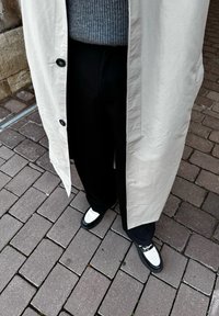 Ανοιχτό μπεζ μακρύ παλτό με μαύρα κουμπιά, γκρι ριγέ πουλόβερ, μαύρα παντελόνια και μαύρα παπούτσια με λευκές λεπτομέρειες σε υφαντή επιφάνεια από κοφτερό κυβόλιθο.