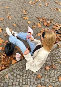 Witte sneakers met bruine strepen, lichtblauwe spijkerbroek, een beige jas, een rood-wit gestreepte voedselkegel vasthoudend, omgeven door herfstbladeren.