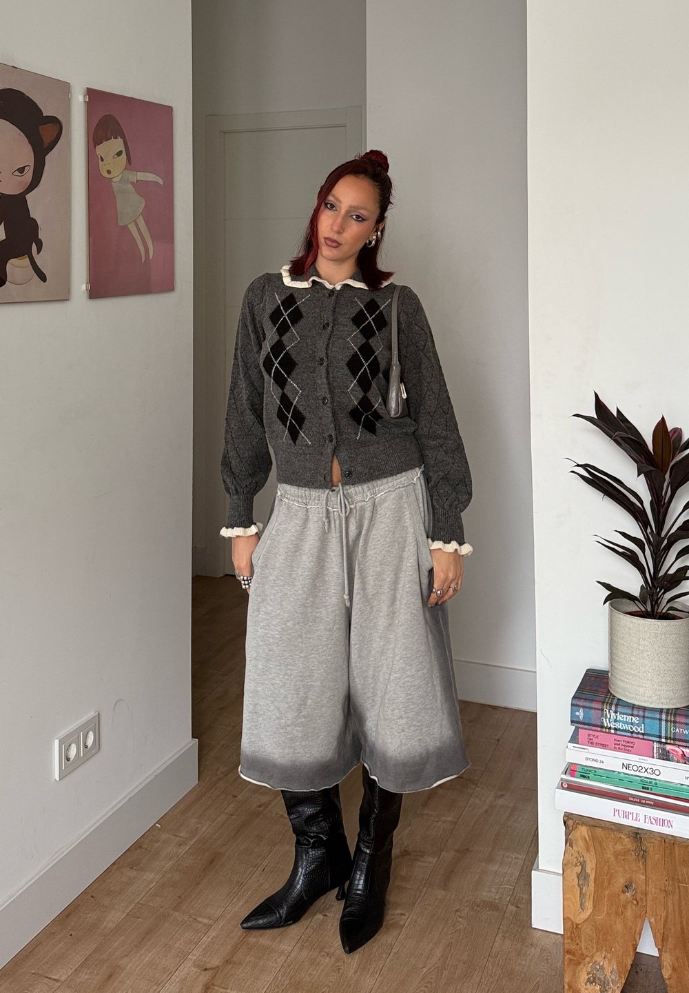 Cardigan en tricot gris à motif losange porté sur un haut blanc à col, associé à un pantalon de survêtement gris ample et des bottes noires montantes jusqu'aux genoux.