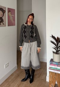 Cardigan en tricot gris à motif losange porté sur un haut blanc à col, associé à un pantalon de survêtement gris ample et des bottes noires montantes jusqu'aux genoux.