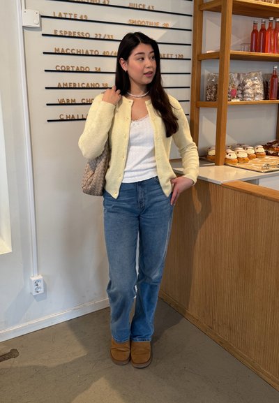 Cardigan amarillo, top blanco sin mangas, jeans azules y botines marrones; bolso trenzado; mostrador de madera y menú de café al fondo.