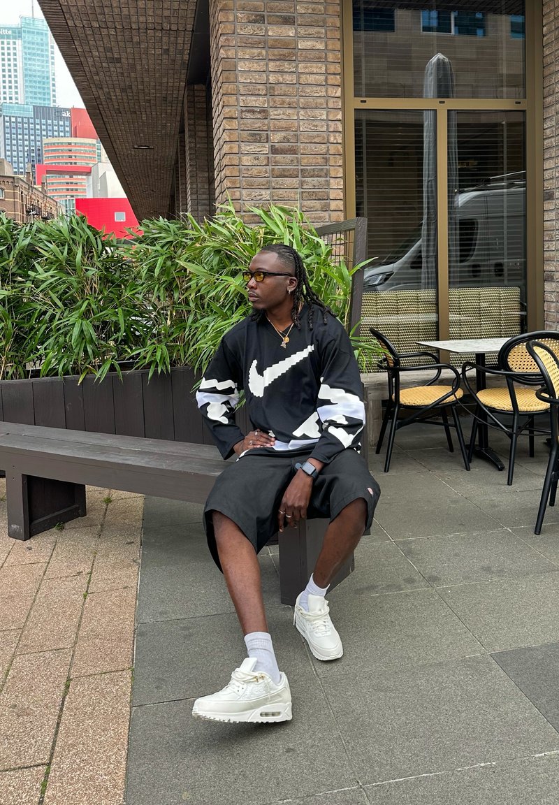 Svart långärmad t-shirt med vitt Nike-logotyp och randiga ärmar, svarta shorts, vita sneakers, en brun trämöbel, grönska i bakgrunden.