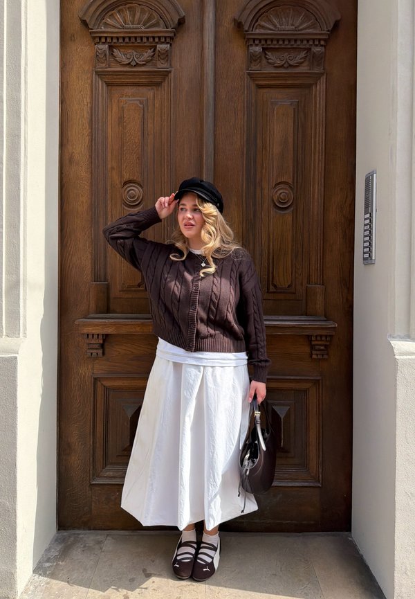 Vrouw in bruine trui, witte rok en zwarte pet staat met een donkere handtas voor een grote gebeeldhouwde houten deur.