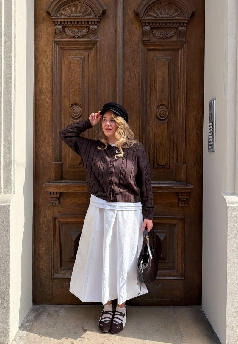 Frau in braunem Pullover, weißem Rock und schwarzer Kappe steht und hält eine dunkle Handtasche vor einer großen geschnitzten Holztür.