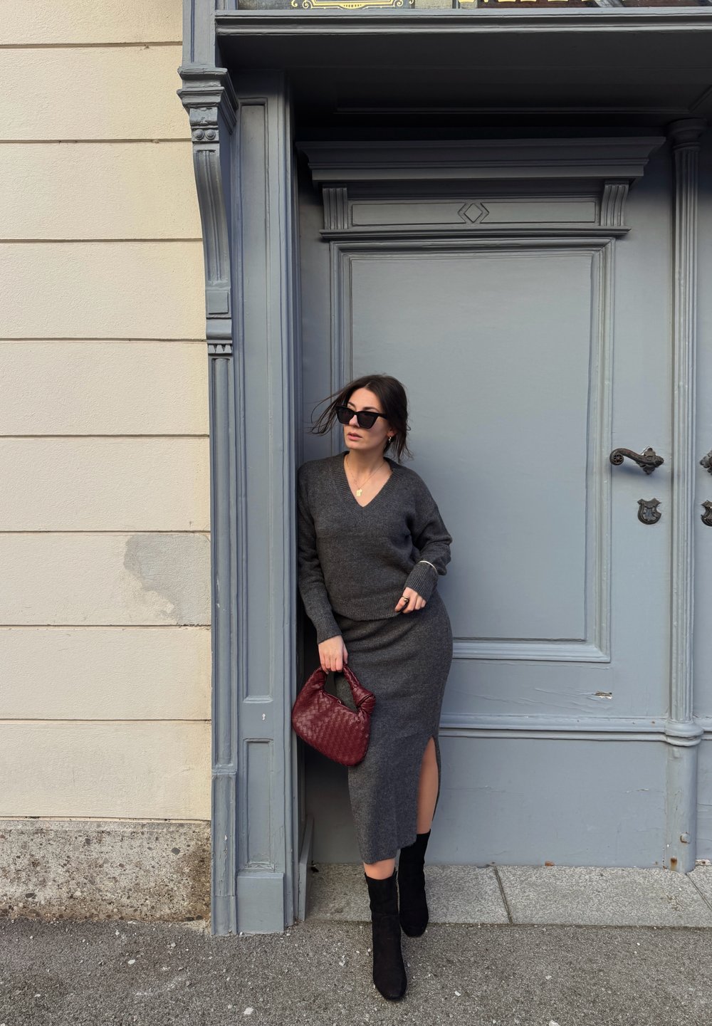 Pull gris en maille et jupe midi assortie avec une fente latérale. Bottes noires jusqu'aux genoux. Tenant un sac à main bordeaux. Debout près d'une porte grise.
