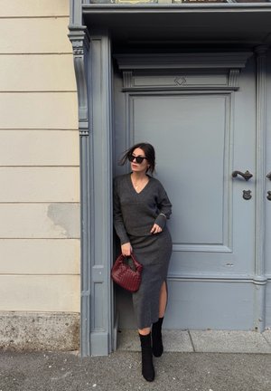 Suéter gris de punto y falda midi a juego con una abertura lateral. Botas negras hasta la rodilla. Sosteniendo un bolso burdeos. De pie junto a una puerta gris.