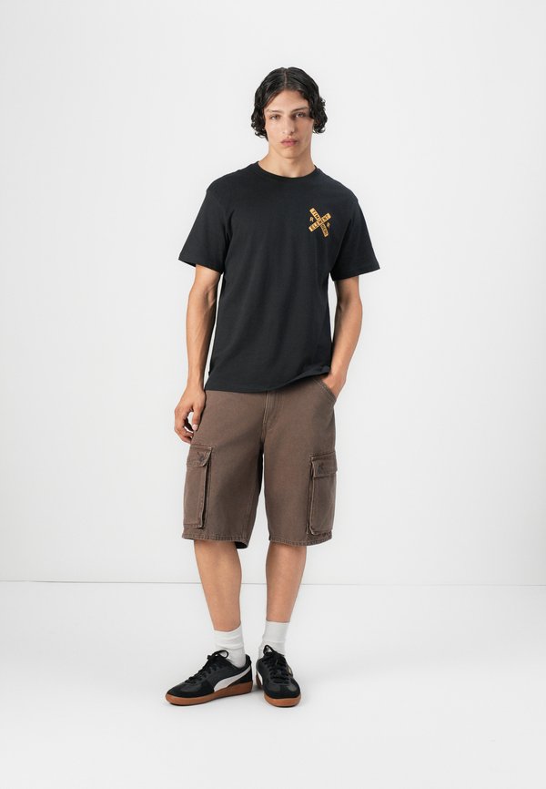 Fekete pamut póló sárga grafikával a mellkason, barna cargo sorttal párosítva, amely két elülső zsebet és laza fazont kínál. Fekete sportcipő.