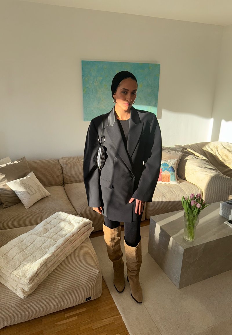Femme portant un blazer noir oversize, des bottes beige montant jusqu'aux genoux, un foulard noir, se tenant dans un salon lumineux avec un canapé et des tulipes sur une table.