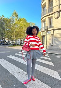 Röd och vit randig tröja, grå plisserad kjol, grå strumpbyxor, skor med röda accenter och en rosa axelväska, med en urban bakgrund.