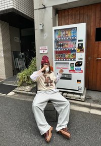 Personne en tenue décontractée assise sur une chaise, buvant dans une tasse, à côté d'un distributeur automatique de boissons dans une rue de la ville.