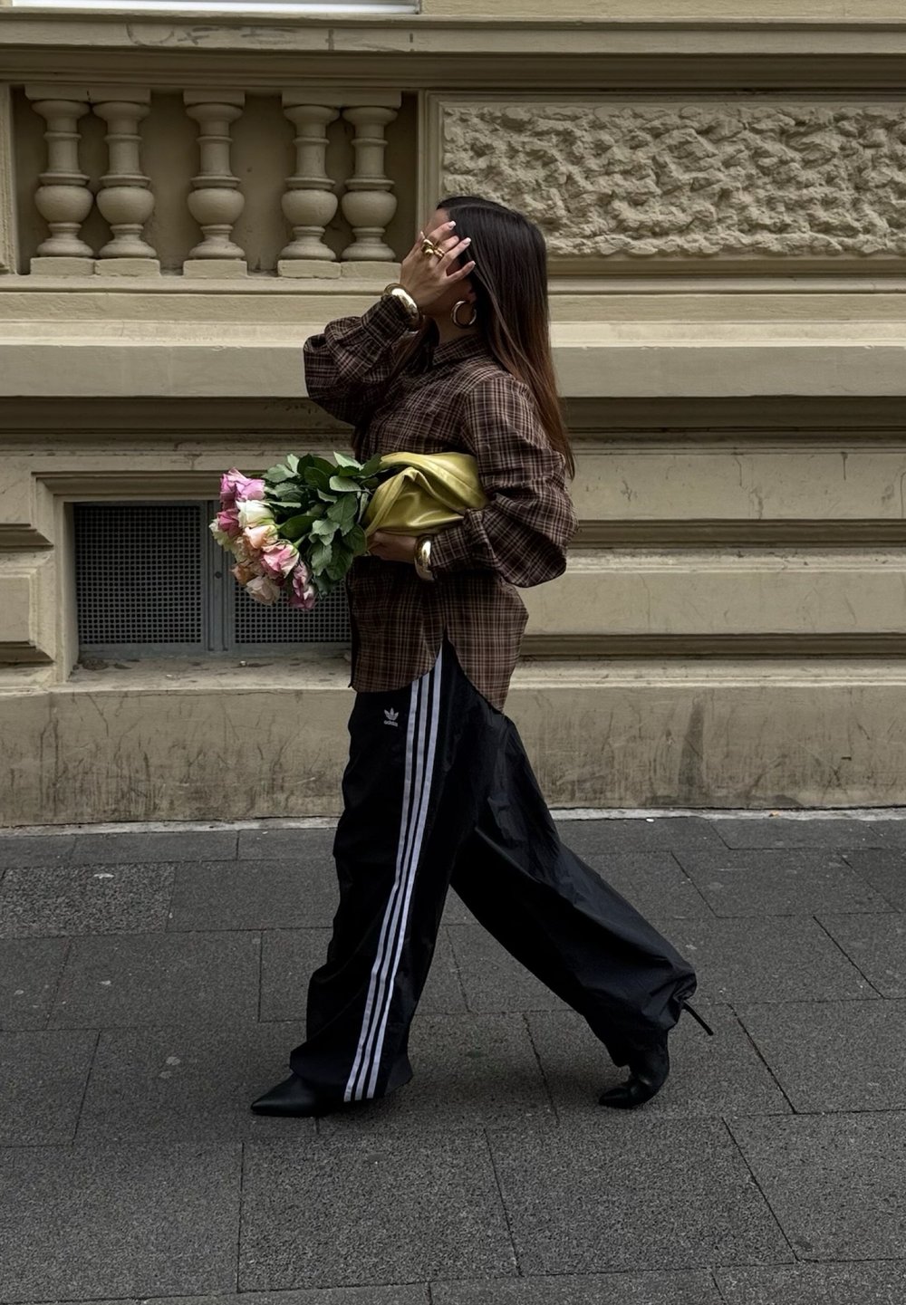 Rjava karirana oversize srajca, črne široke hlače z belimi črtami, črne pete, v rokah drži šopek rož različnih odtenkov roza in bele, zavit v rumeno tkanino.