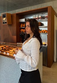 Bluza albă, texturată, cu mâneci clopot, purtată peste o fustă neagră. În fundal, o vitrină de brutărie cu diverse produse de patiserie și un decor din lemn.